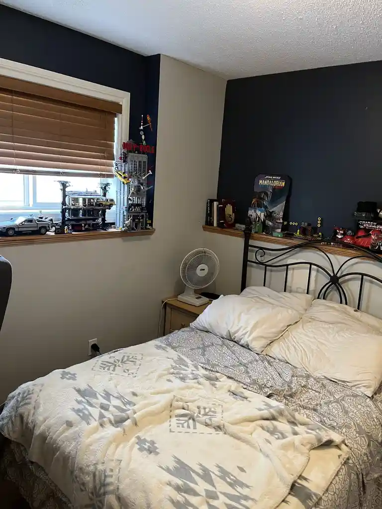 A messy bedroom with an unmade bed, dark walls, and a window sill cluttered with LEGO sets, action figures, and a DeLorean…