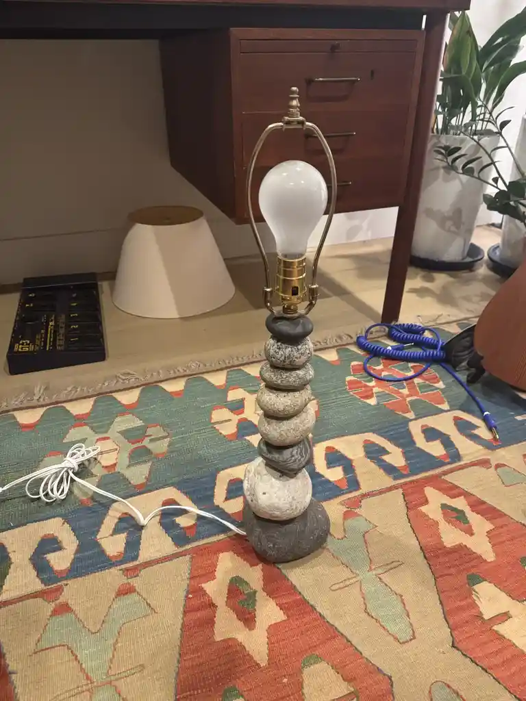 A lamp with a stack of smooth river rocks as its base sits on a colorful patterned rug, its white bulb unlit. The lamp's…