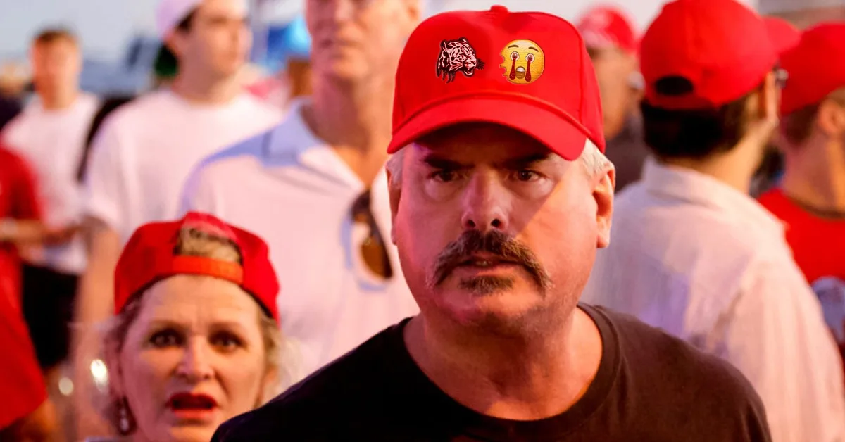 A man with a mustache wearing a red cap with a jaguar logo and a crying emoji on it stares intensely forward. Behind him, a…