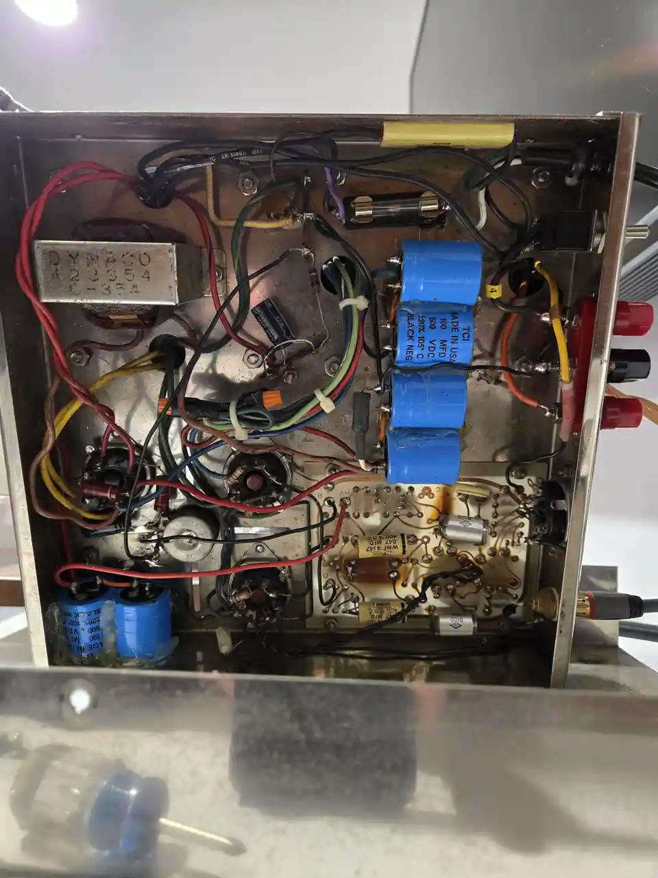 The image shows the interior of an old, open electronic amplifier with visible wiring, capacitors, tubes, and circuit…