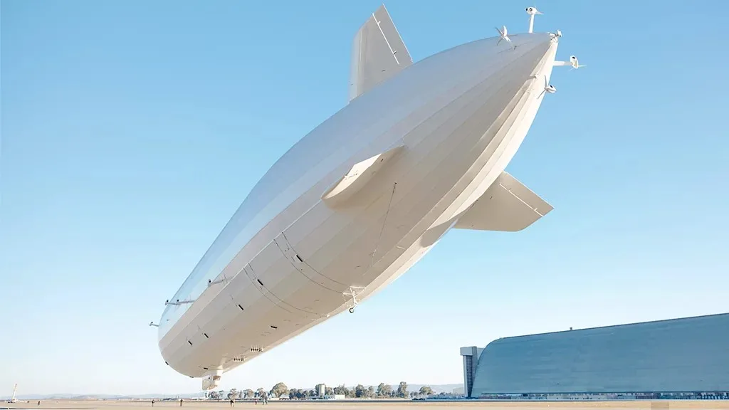 A large, white, modern-looking airship, resembling a zeppelin, is shown tilted at an angle against a clear blue sky…