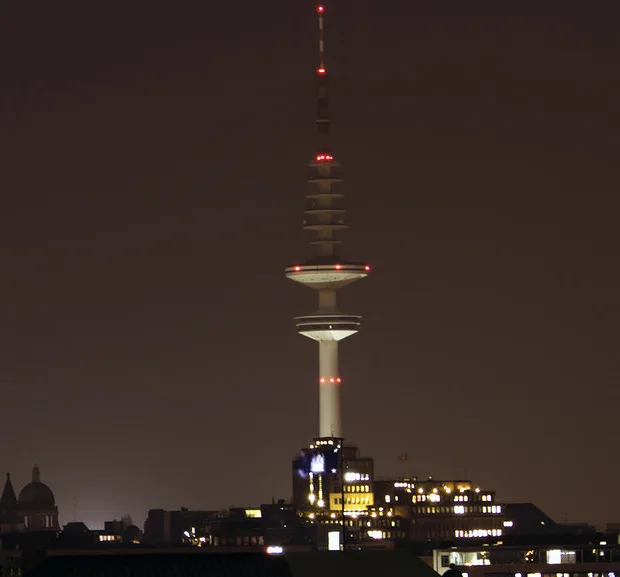 A nighttime photo of a tall, illuminated tower with red lights against a dark sky, with city buildings visible below. The…