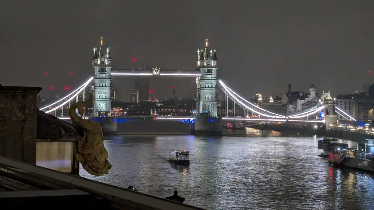 A nighttime photo of Tower Bridge in London, illuminated with white and blue lights, spanning the River Thames with a boat…