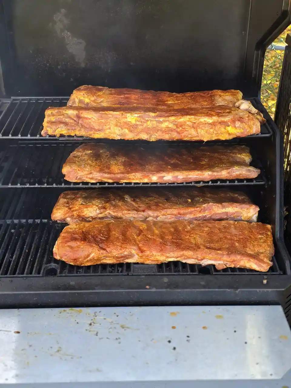 Five racks of seasoned ribs are stacked on the grates of a black outdoor grill, with smoke rising from the meat. The text…
