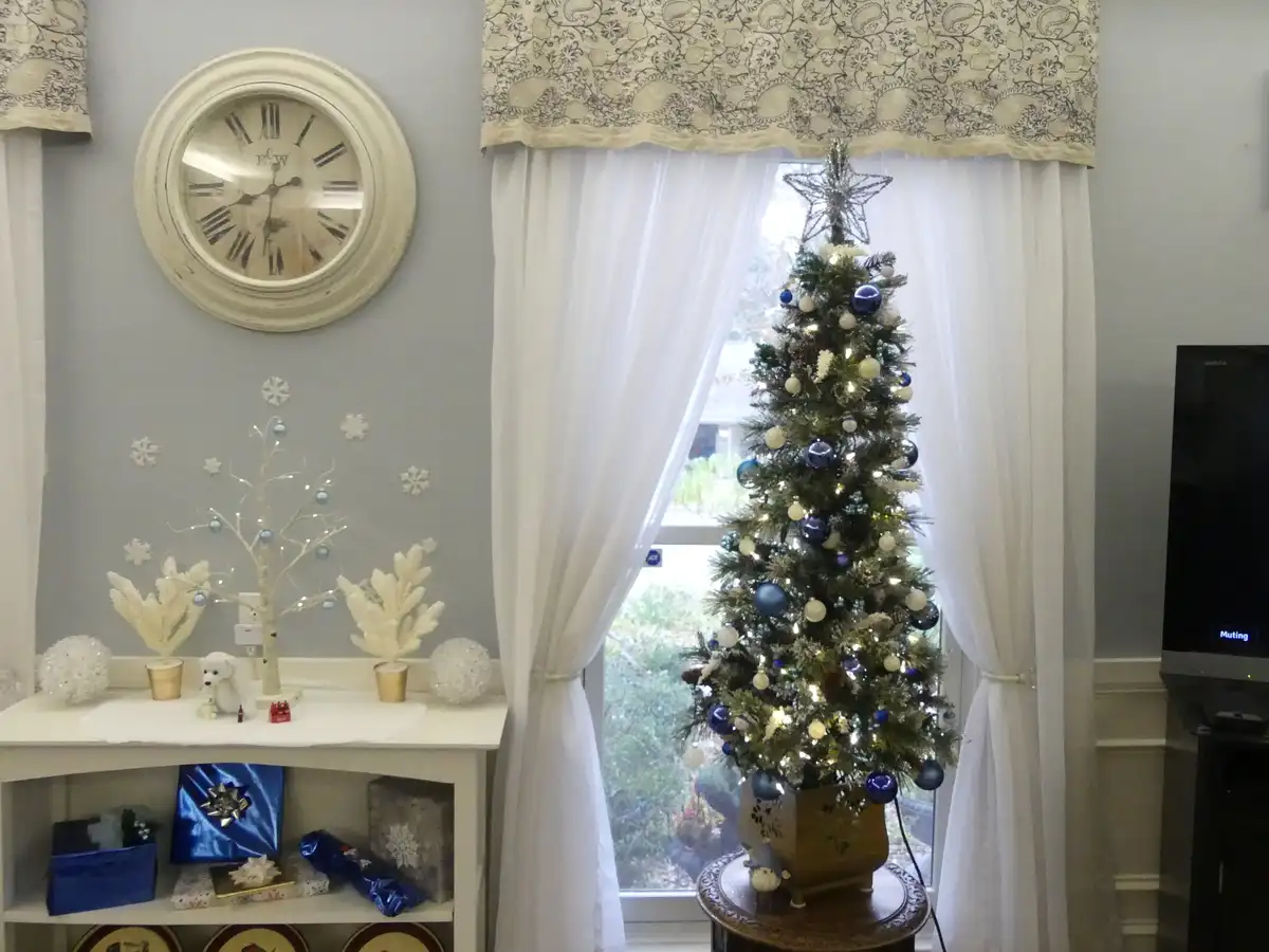 A decorated christmas tree with blue and white ornaments and a star on top stands in front of a window with white curtains…