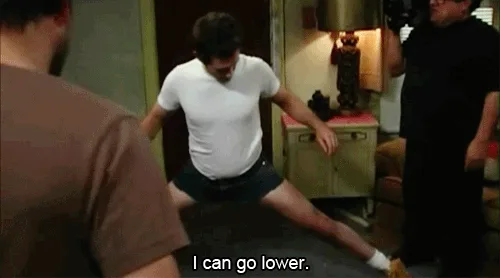 A man in a white t-shirt and dark shorts performs a wide-legged split on a rug in a room, repeating the phrase 'I can go…
