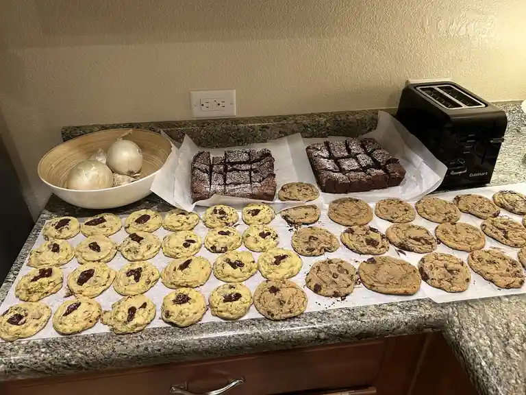 A kitchen counter displays a large batch of chocolate chip cookies, two trays of brownies, and a bowl of onions, with…