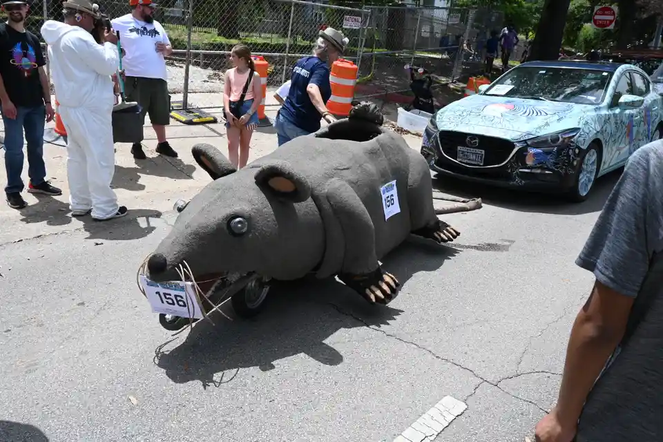 A large, gray rat-shaped vehicle with the number 156 on its mouth and side is parked on a street during what appears to be a…