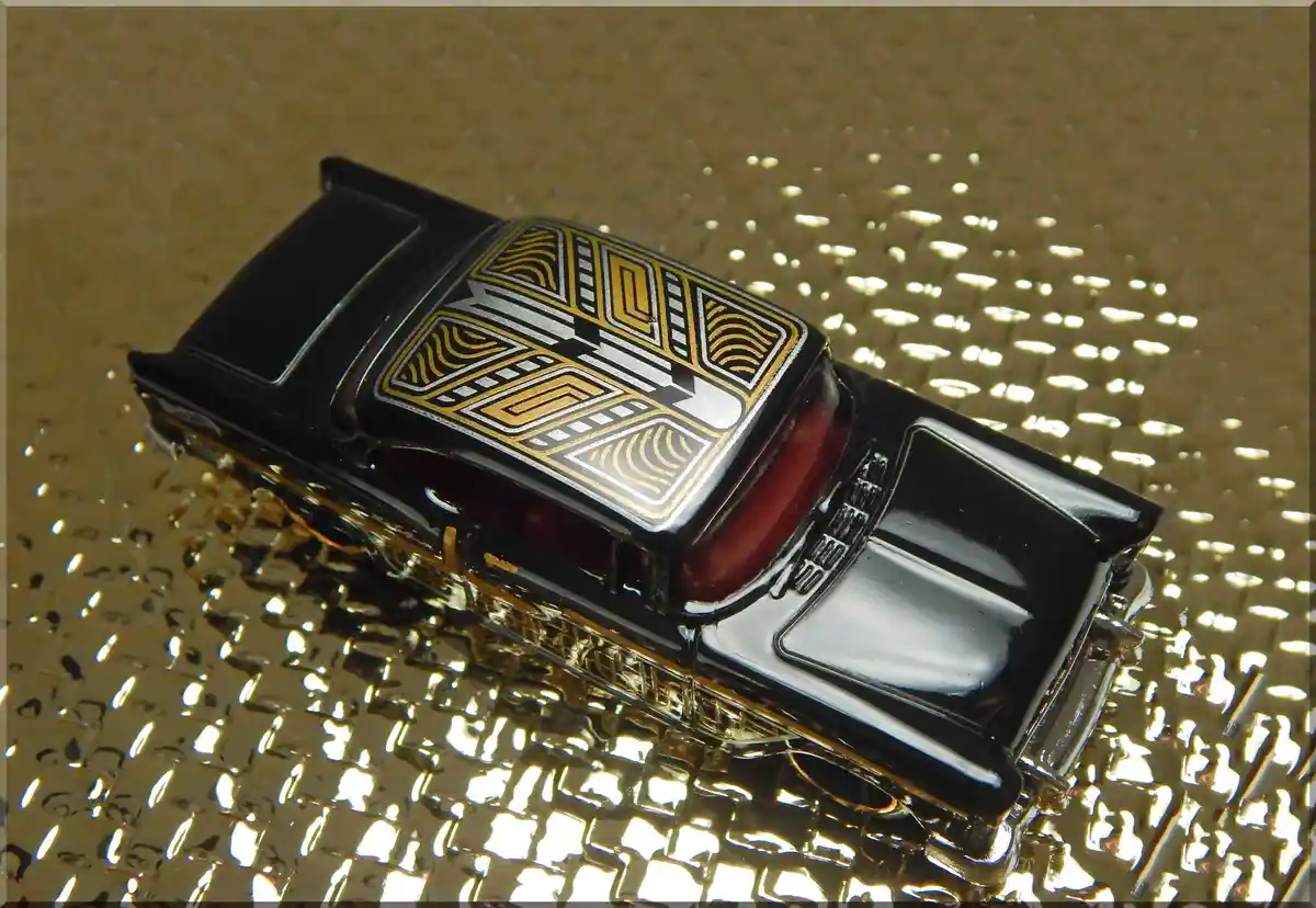 A black toy car with a gold and silver geometric pattern on its roof sits on a textured gold surface. The overlaid text…