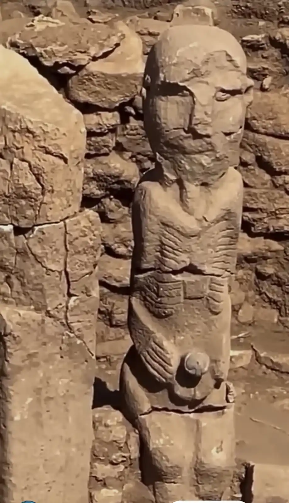 A weathered ancient stone statue with a carved face and torso stands in front of a stone wall, its right eye socket missing…