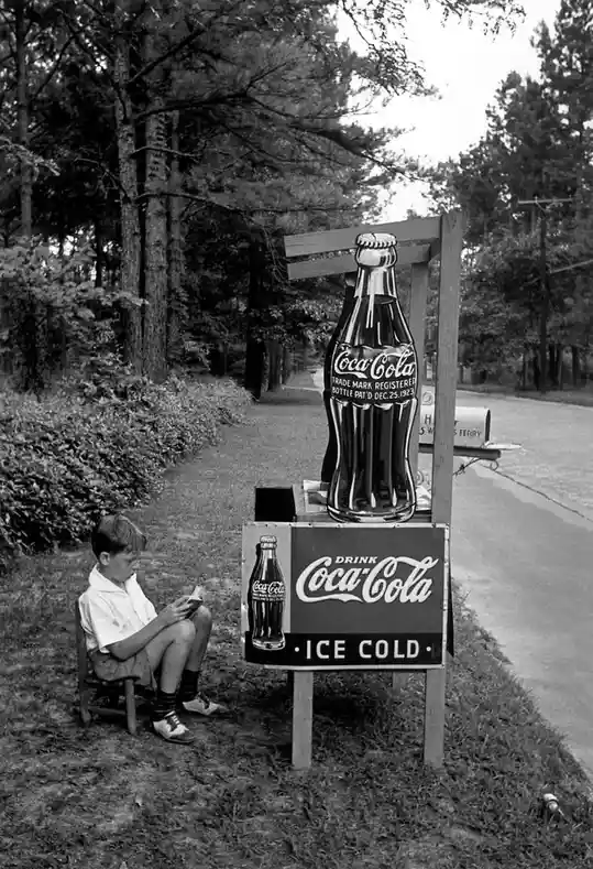 A vintage black-and-white photo shows a young boy sitting on a small wooden chair beside a roadside Coca-Cola advertisement…