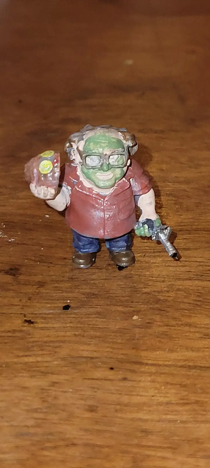 A hand-painted miniature figurine of an elderly man with green skin, glasses, and a red shirt stands on a wooden surface. He…