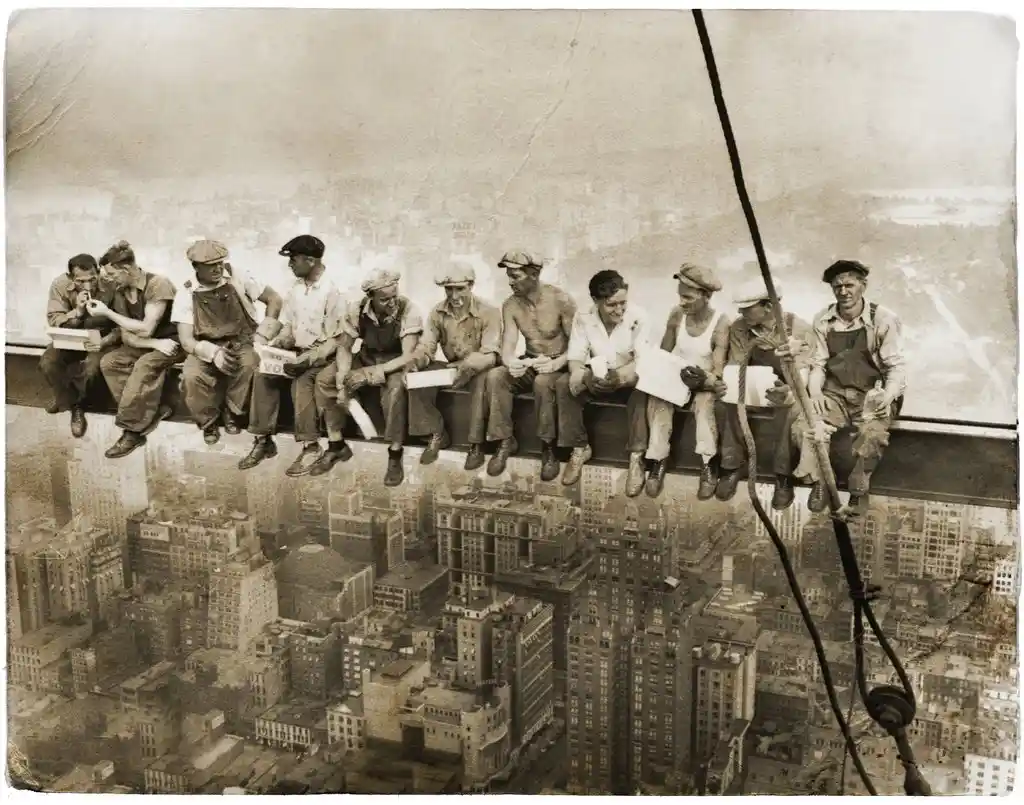 A sepia-toned historical photograph shows ten construction workers sitting casually on a narrow steel beam high above a…
