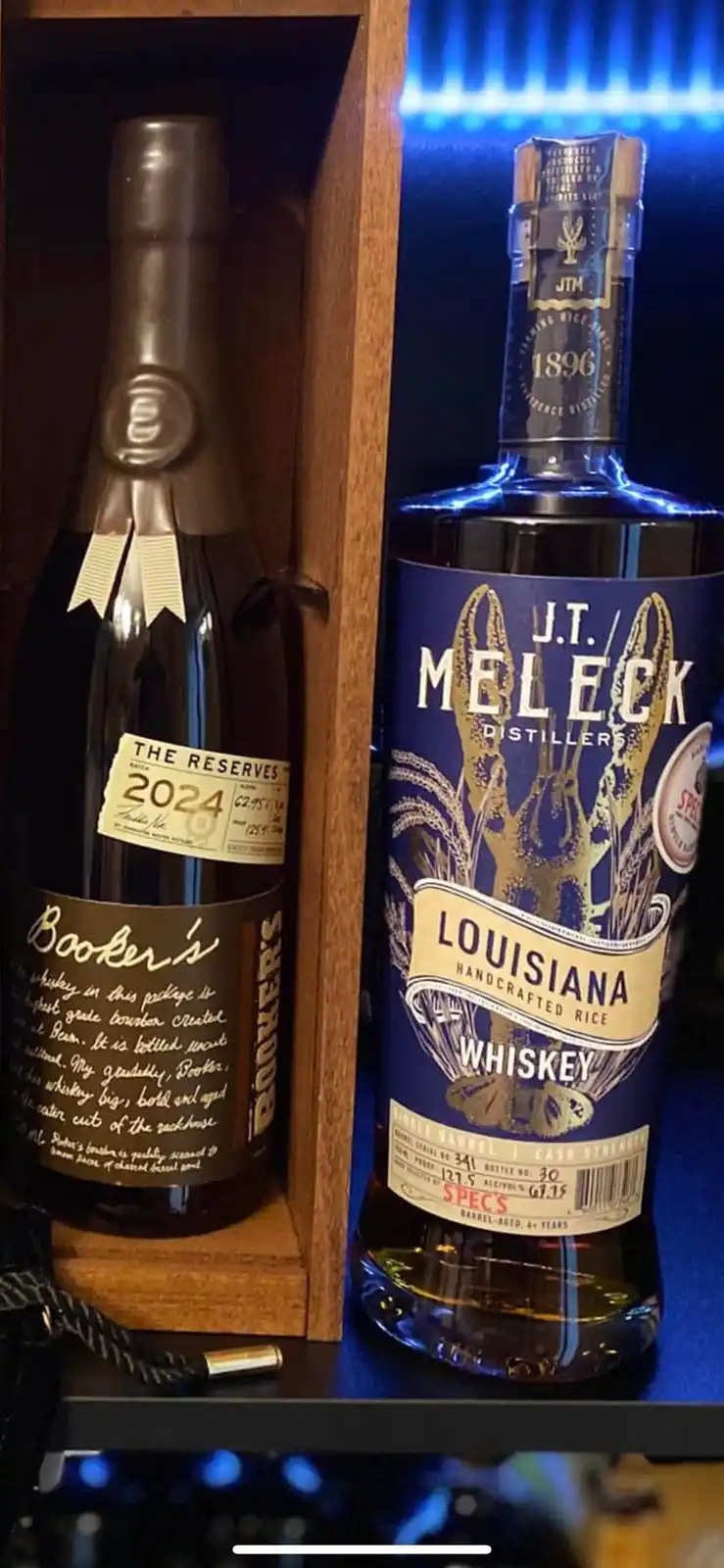 Two whiskey bottles sit side-by-side on a shelf: a Booker's 2024 bourbon in a wooden box and a J.T. Meleck Louisiana…