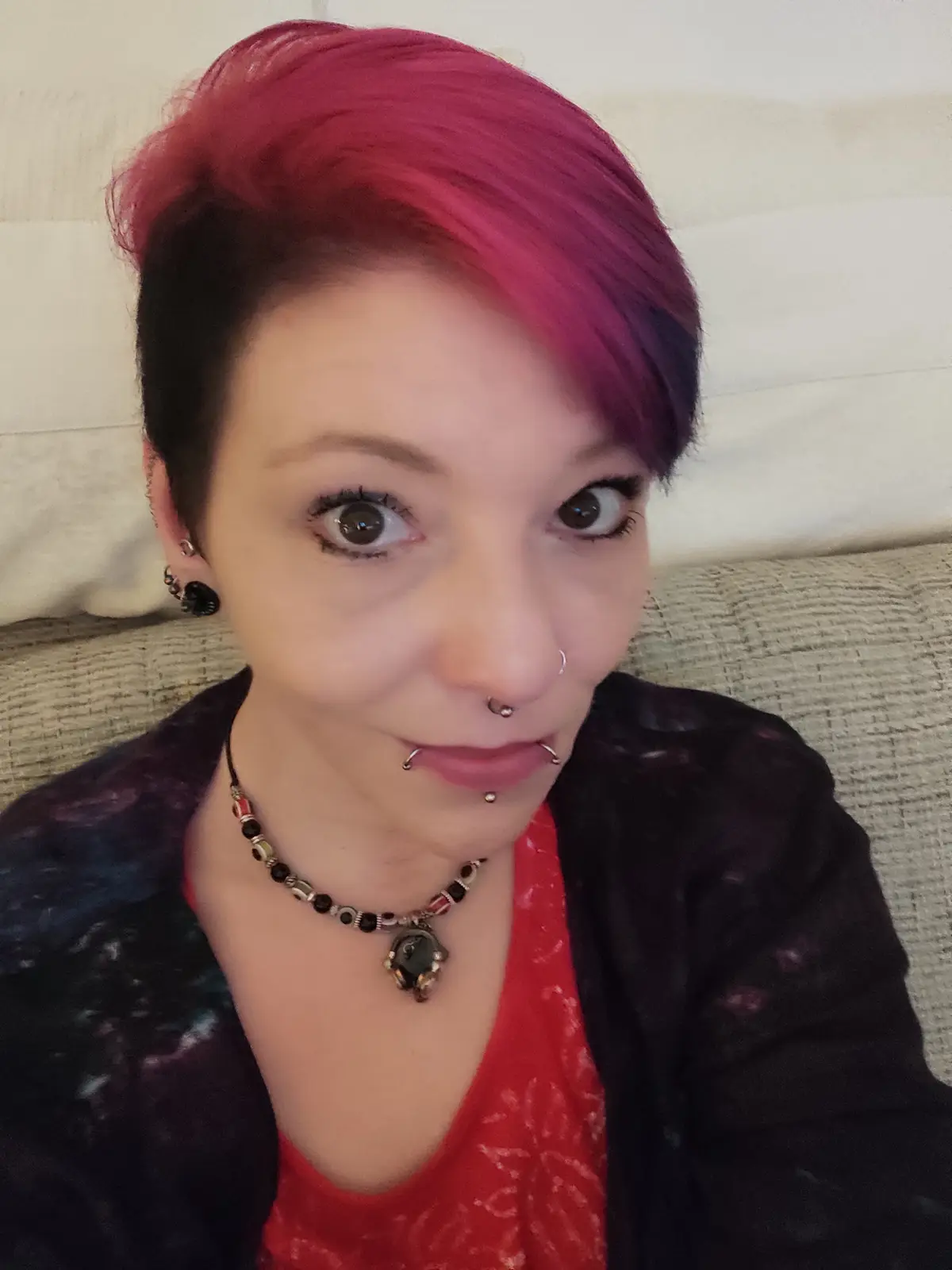 A person with short, brightly dyed magenta and purple hair sits on a couch, looking directly at the camera with wide eyes…