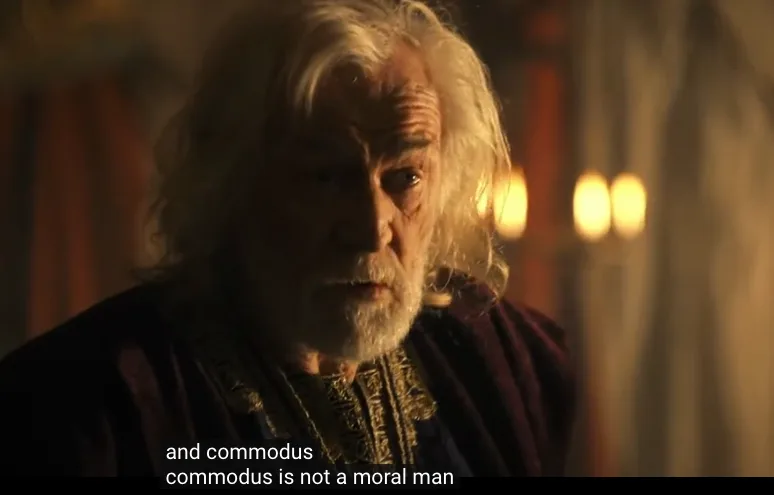 A close-up shot of an elderly man with long white hair and a beard, wearing ornate dark robes, looking intently forward in a…