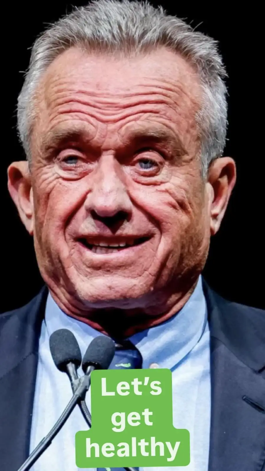 A close-up photo of an older man with gray hair, wearing a suit and tie, smiling while standing behind two microphones. A…