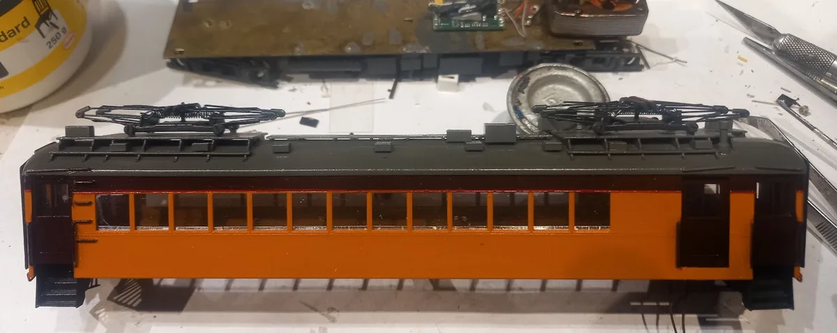 A close-up photo of a detailed model train car with an orange lower body and dark upper section, featuring windows and…