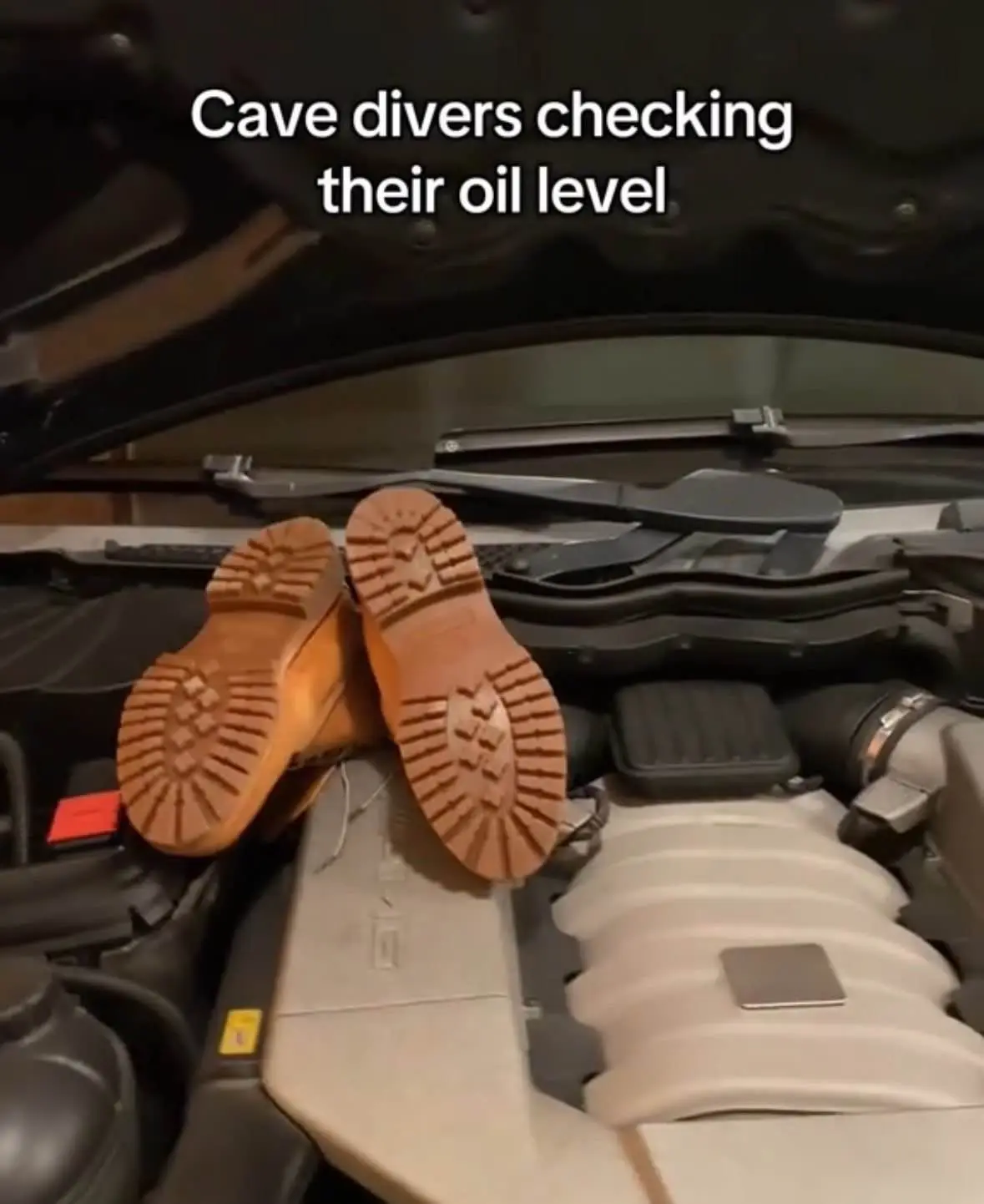 A pair of brown work boots with deep tread soles are placed upside down on top of an open car engine, with the text 'Cave…