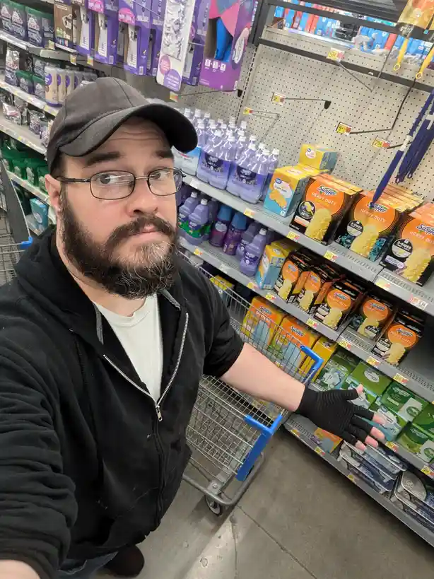 A man with a beard, glasses, and a cap takes a selfie in a store aisle, gesturing with his gloved hand toward shelves…