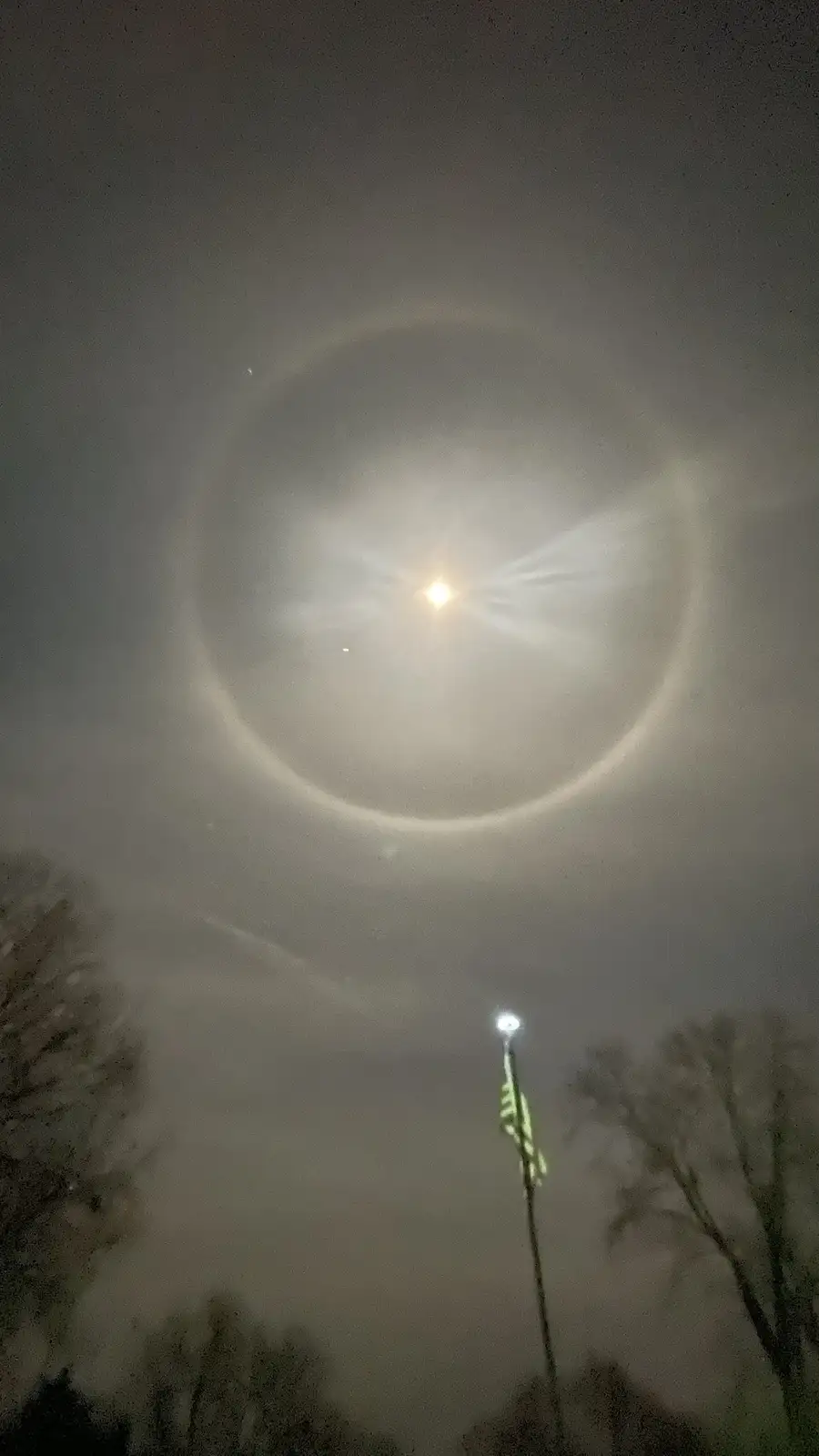 A night sky photo shows a bright moon with a distinct, glowing circular halo around it, set against a hazy, gray sky…