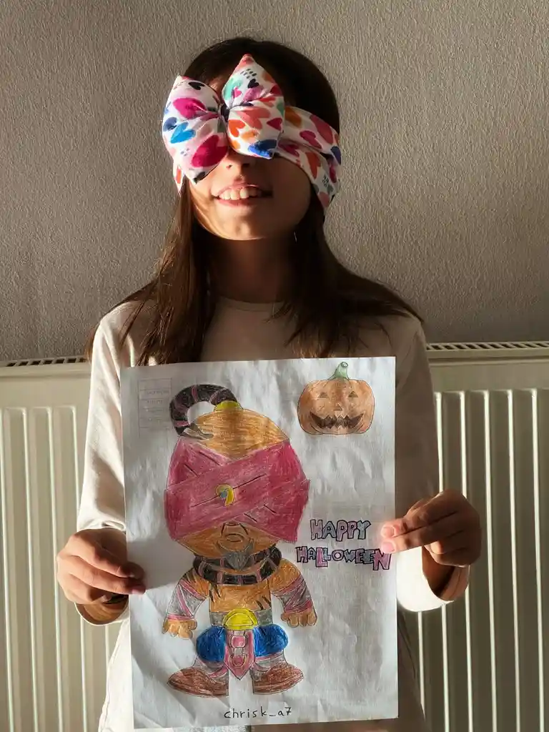 A young girl with a large, colorful bow blindfold covering her eyes smiles while holding up a child's Halloween drawing. The…