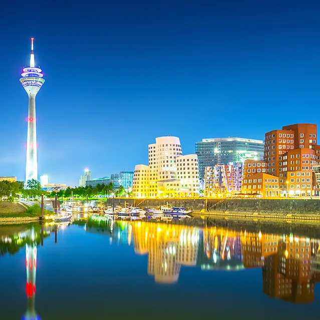 A vibrant nighttime cityscape featuring a tall, illuminated tower with colorful lights and a modern skyline reflected in a…