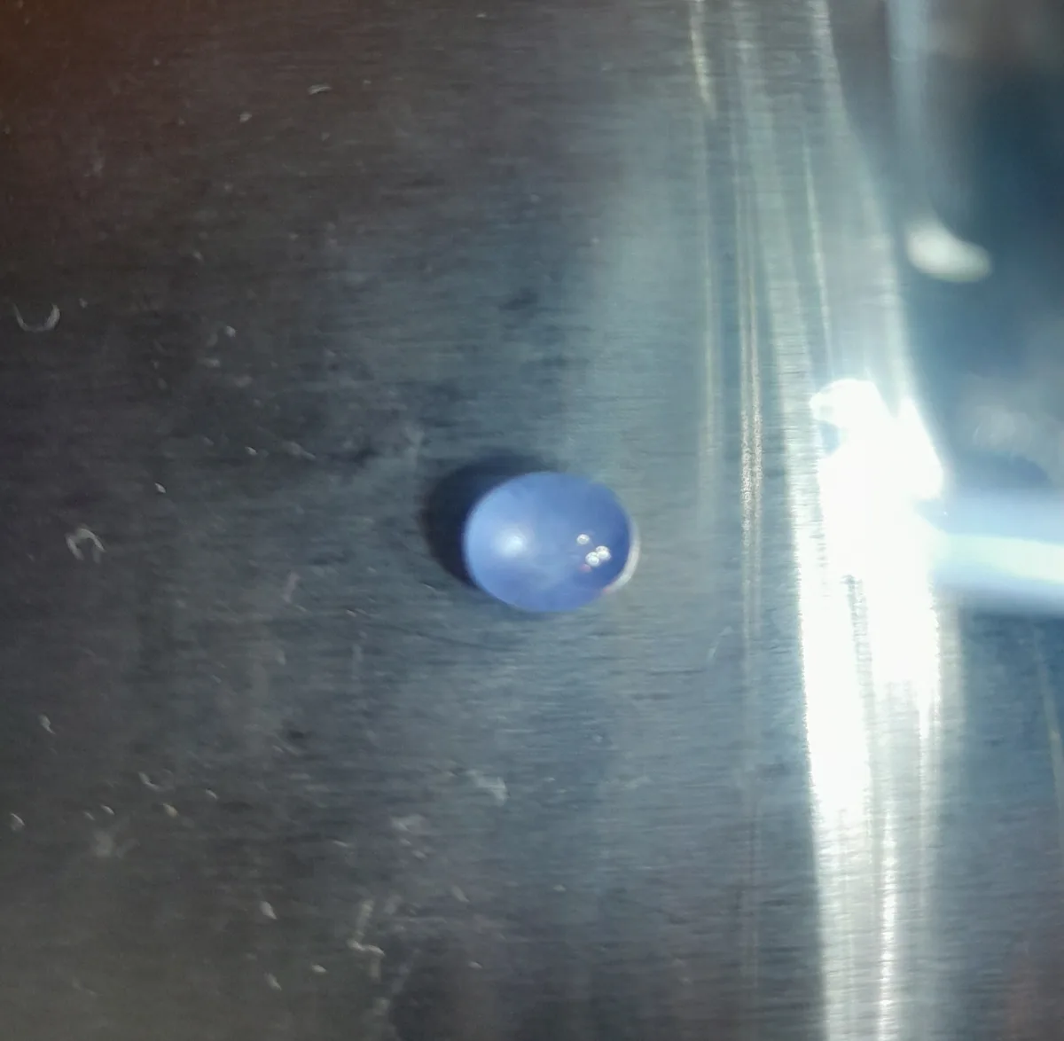 A small, round, blue object resembling a blueberry sits on a brushed metal surface with visible scratches and bright light…