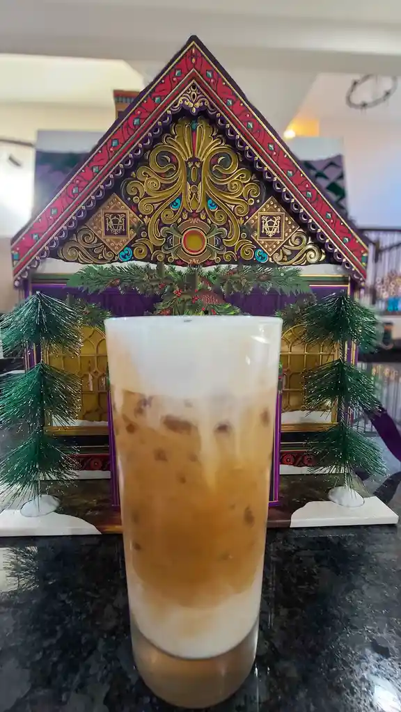 A tall glass of iced coffee with a creamy top sits on a dark countertop, positioned in front of an ornate, colorful…