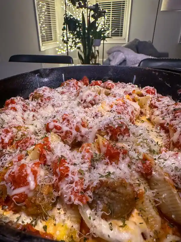 A close-up photo of a skillet filled with pasta shells, meatballs, tomato sauce, melted cheese, and grated parmesan cheese…