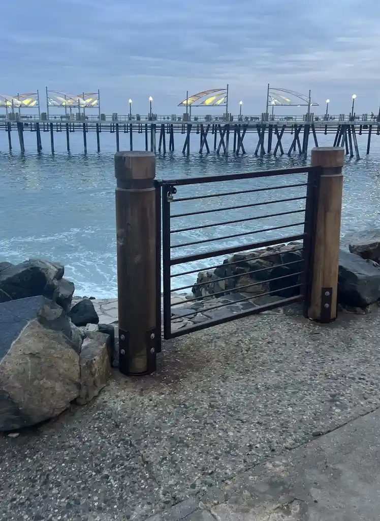 A photo shows a small metal gate with wooden posts, locked with a padlock, blocking access to a concrete path leading to a…