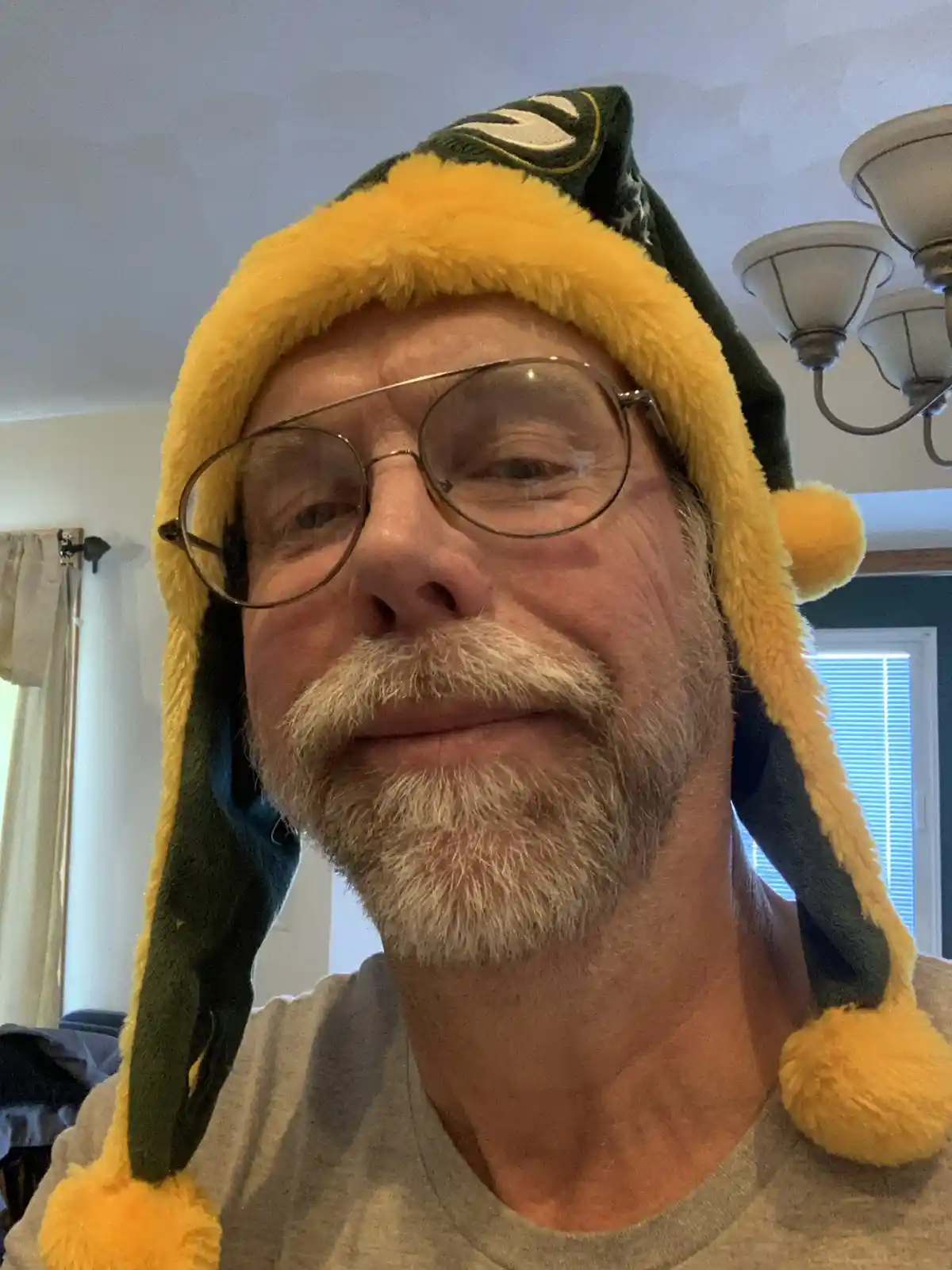 A middle-aged man with glasses and a gray beard wears a green and yellow Green Bay Packers winter hat with ear flaps and…