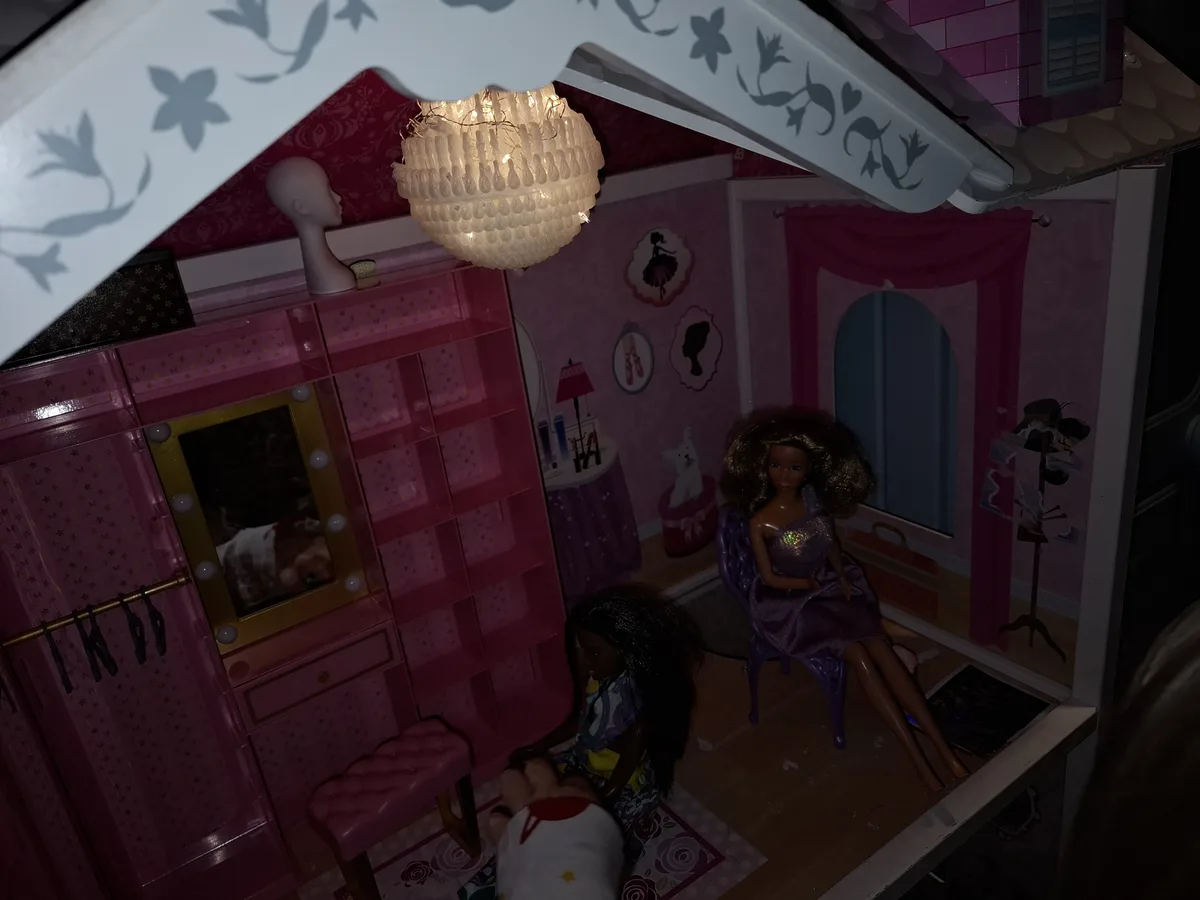 A dollhouse bedroom with pink walls, a chandelier, and two dolls — one seated in a purple dress, the other kneeling. A real…
