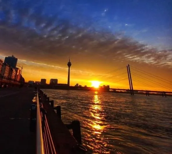 A scenic sunset over a river with a city skyline, featuring a prominent tower and a suspension bridge. The sun is low on the…