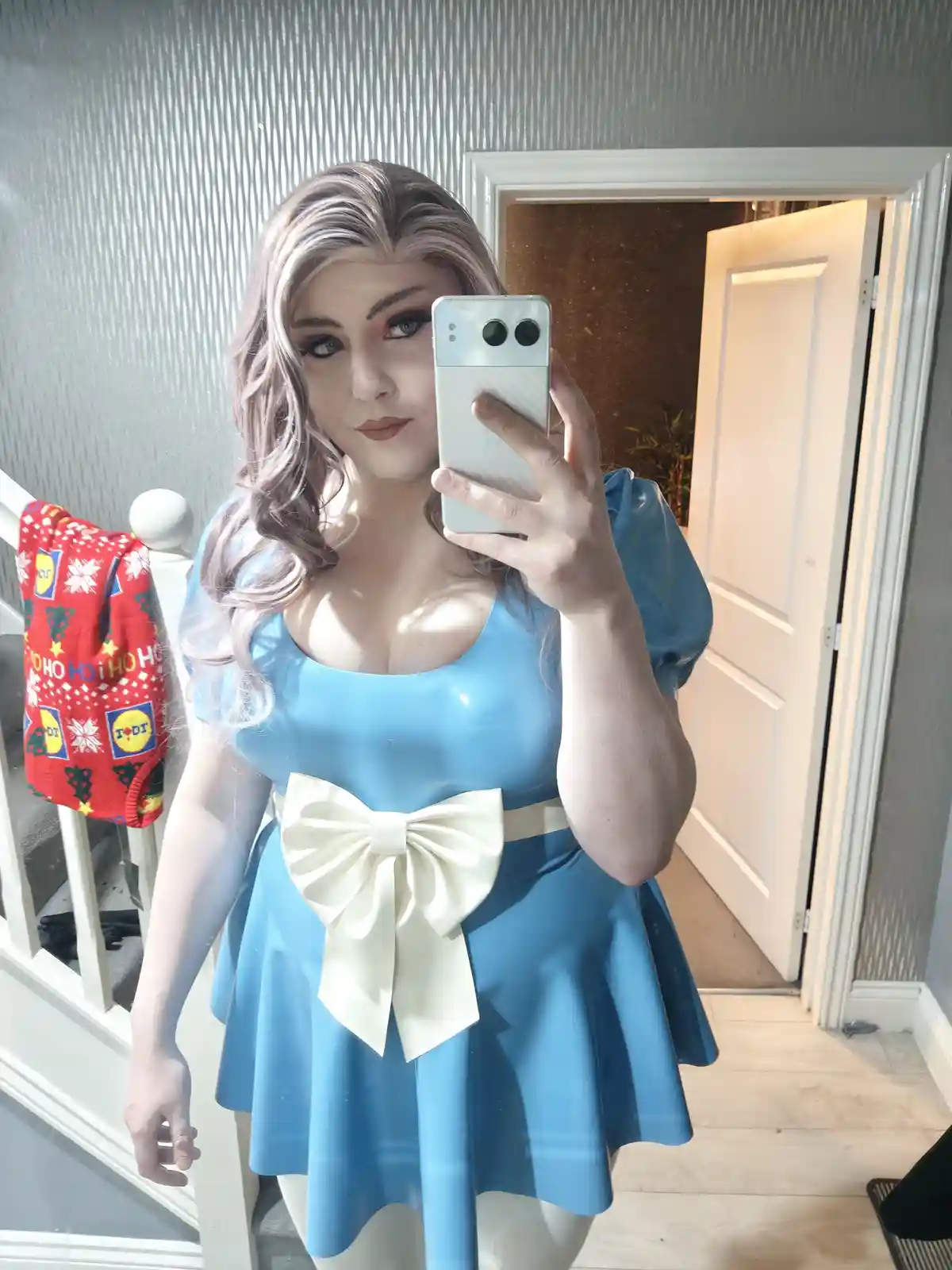 A person in a blue dress with a large white bow and light purple hair takes a mirror selfie holding a white phone. Behind…