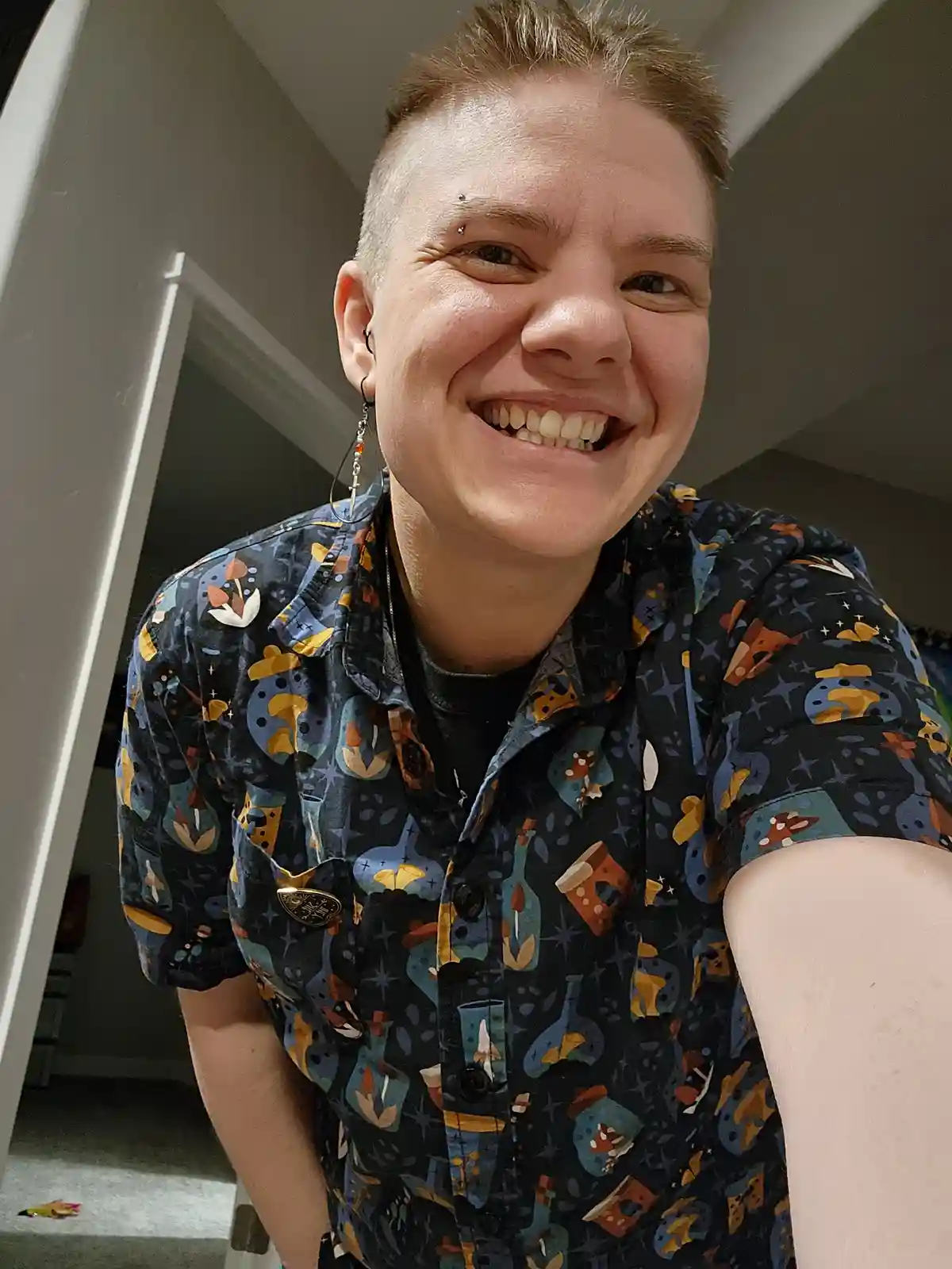 A person with short hair and multiple facial piercings smiles broadly at the camera while wearing a dark, patterned…