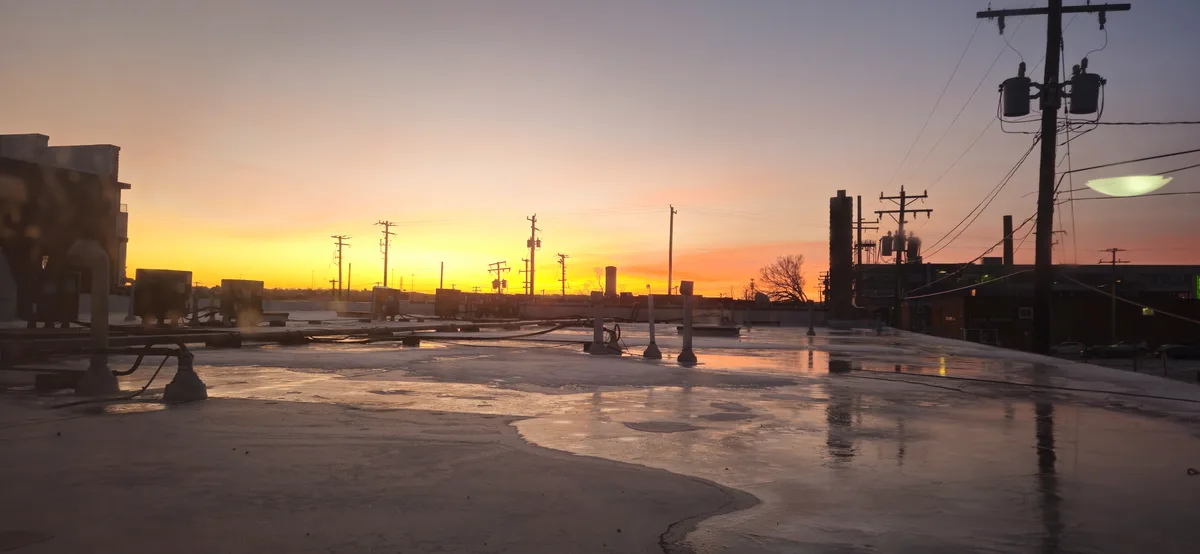 A rooftop at sunrise with wet patches reflecting the orange and yellow sky, surrounded by utility poles and industrial…