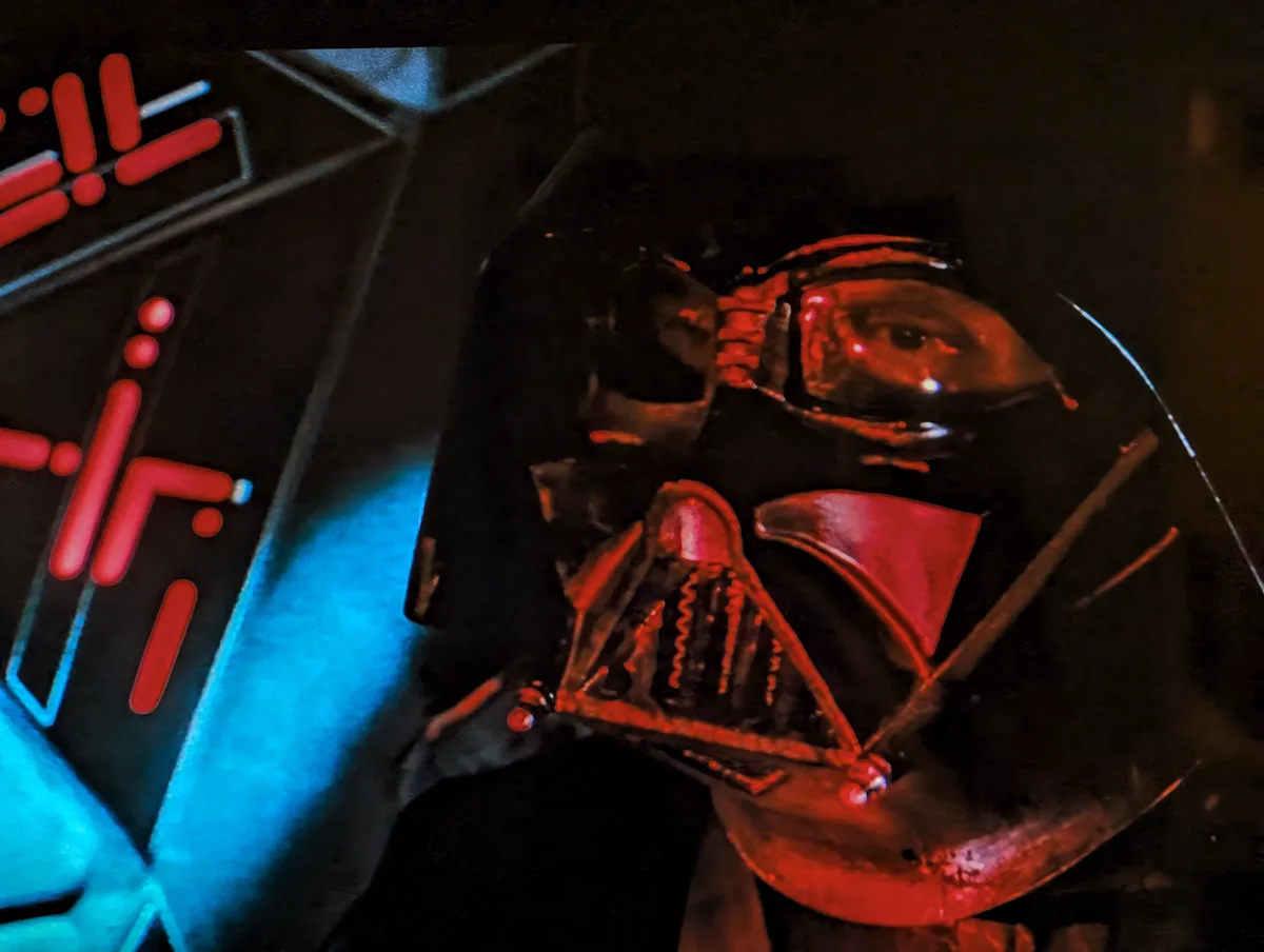 A close-up photo shows the helmet of Darth Vader, illuminated by red and blue lights, with a portion of a glowing red and…