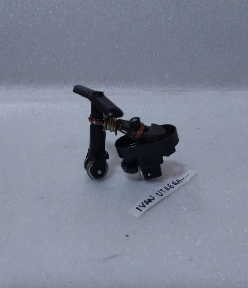 A small, black, broken mechanical component with wheels and exposed copper wire sits on white foam. A handwritten label…