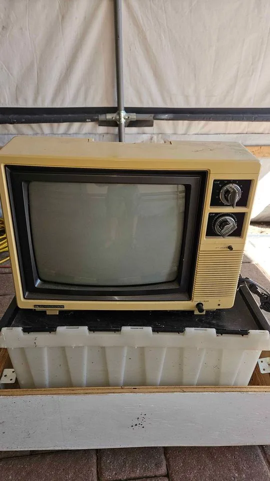 A vintage beige television set with two dials on the right side sits atop a white plastic storage bin, placed on a wooden…