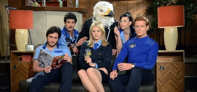 A group of six people, including one wearing a large eagle head costume, are seated on a couch in a retro-style living room…