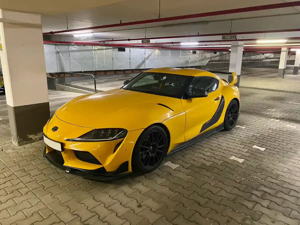 A bright yellow Toyota Supra sports car is parked in an underground parking garage with red pipes and numbered pillars. The…