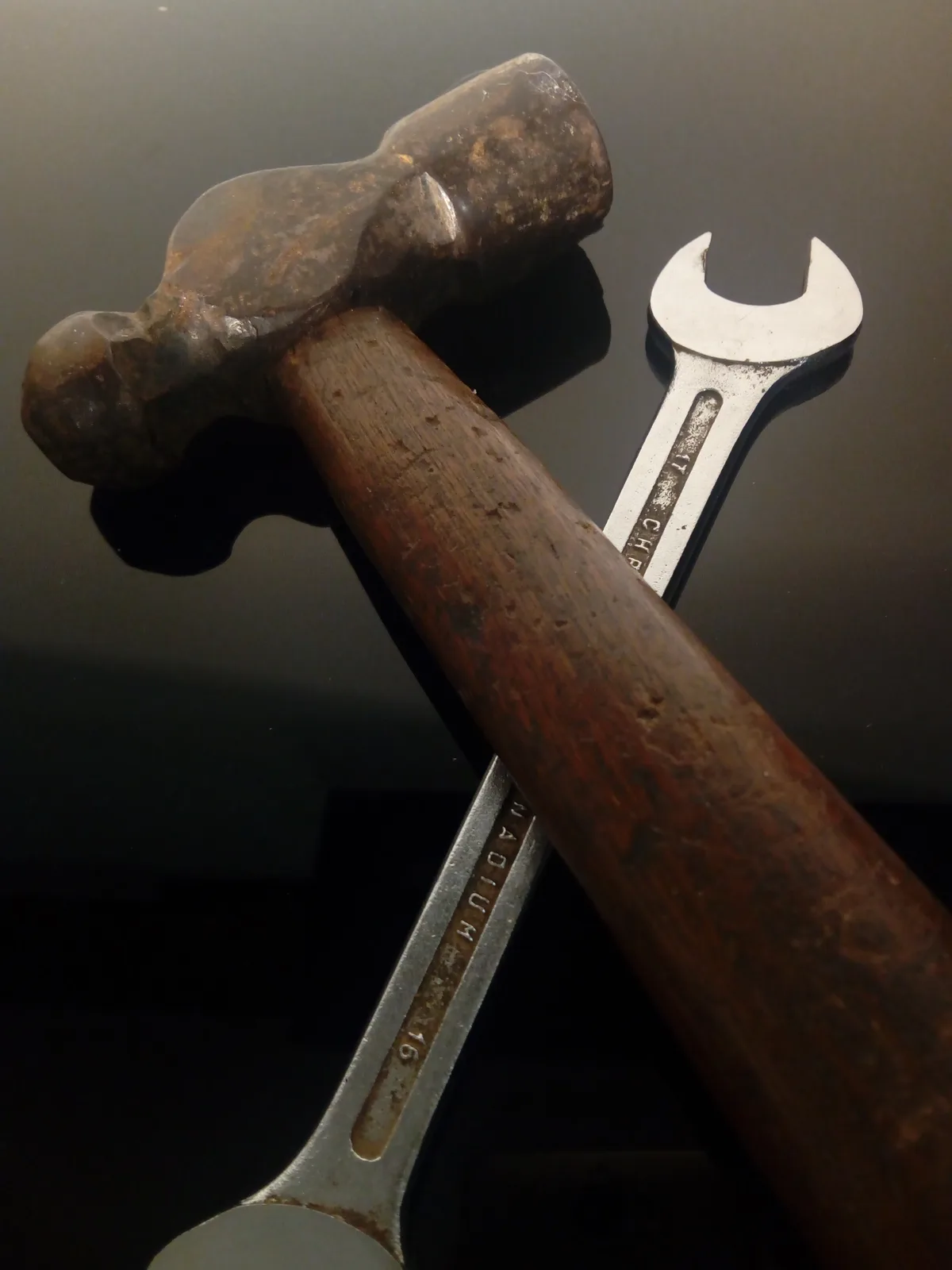 A close-up photo shows a well-worn claw hammer with a wooden handle and a metal wrench lying on a dark, reflective surface…