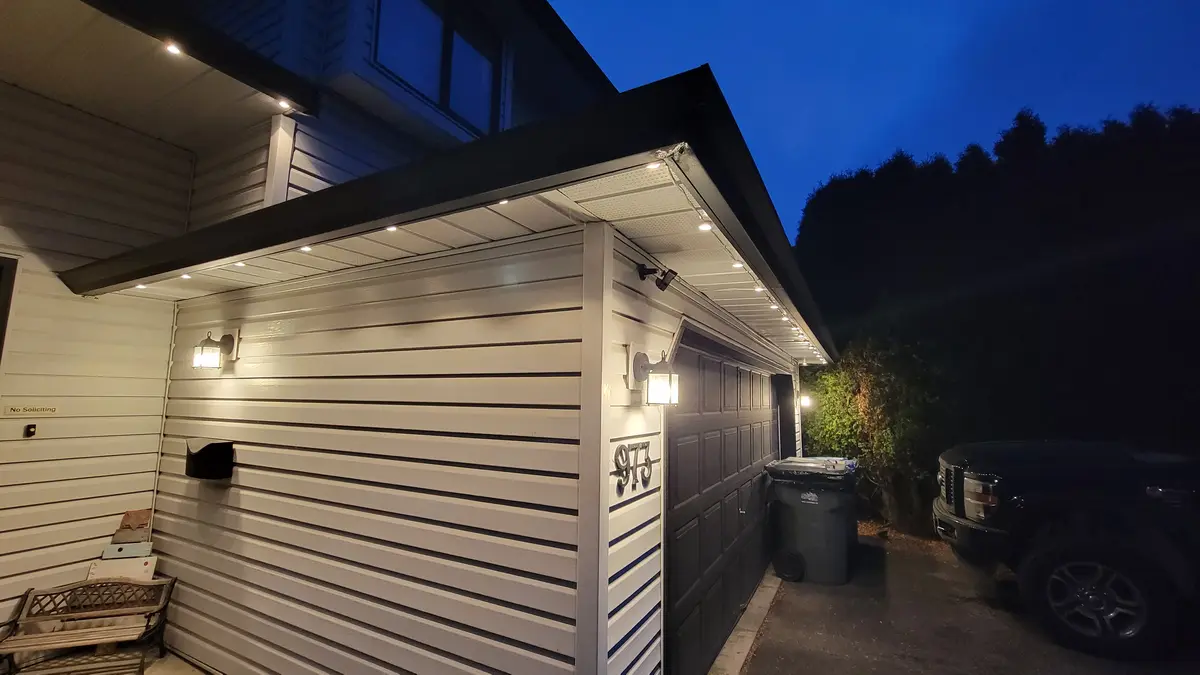 A nighttime photo of a house exterior with white siding, a dark garage door, and a lit porch awning with string lights. A…