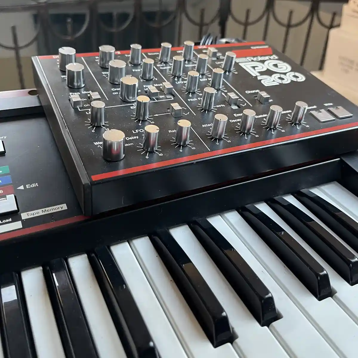 A close-up photo of a Roland PG-200 synthesizer, showing its black panel with numerous silver knobs and switches, resting…