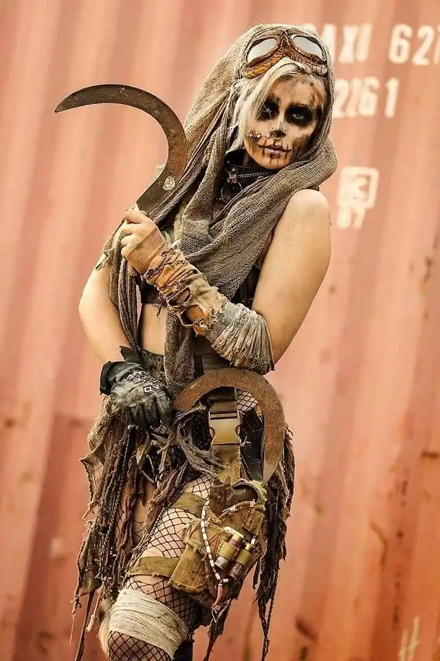 A person dressed in a gritty, post-apocalyptic costume with skull makeup, goggles, and a large curved blade, stands against…