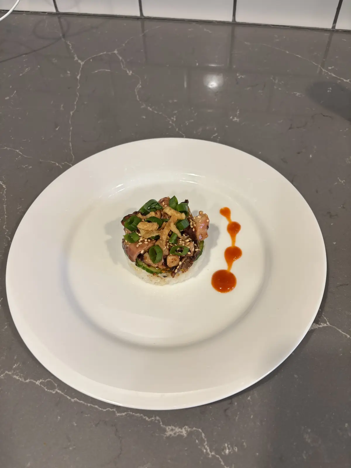 A small, meticulously plated dish sits on a white plate atop a grey marble countertop. The dish consists of a small mound of…