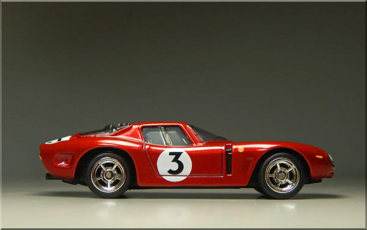 A red die-cast model of a classic race car, shown in profile against a plain gray background, features a large white circle…