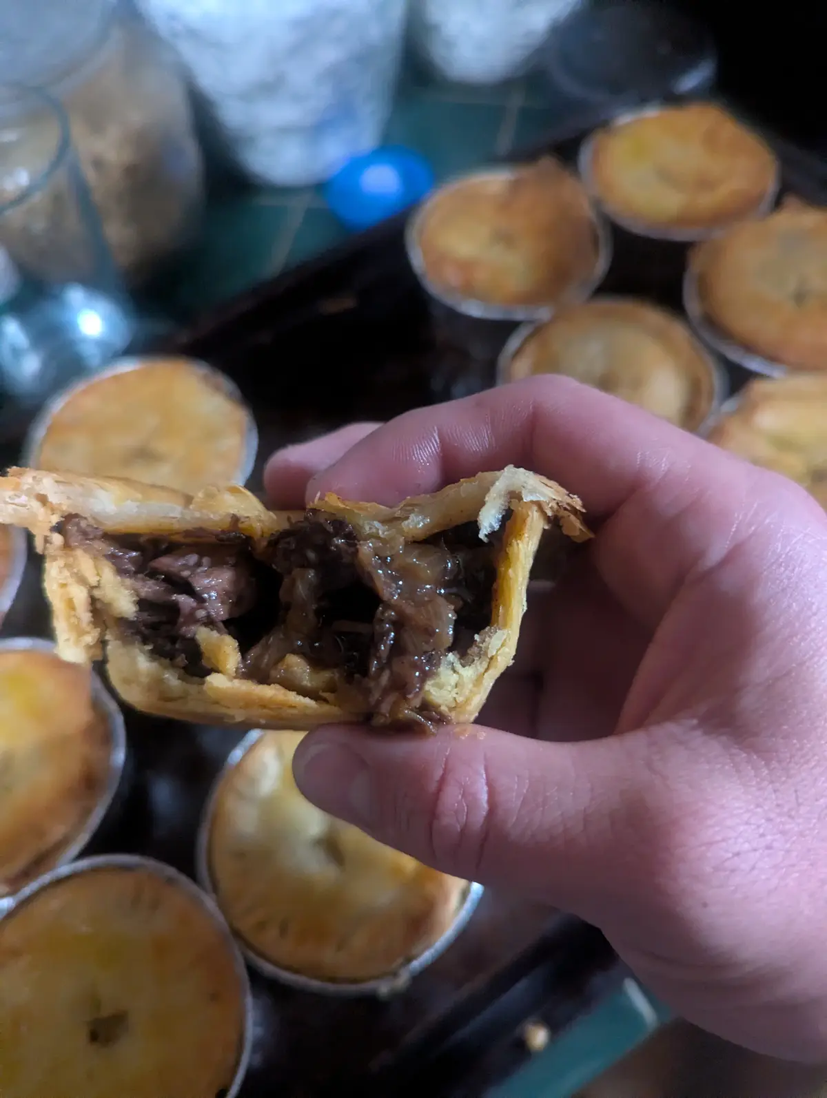 A hand holds a partially eaten beef pie, revealing its savory meat and gravy filling, with several other similar pies in…