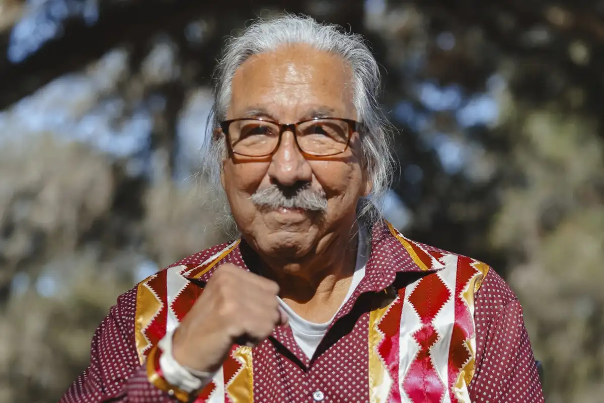 An older man with glasses and a mustache, wearing a colorful patterned shirt, points his finger forward with a confident…